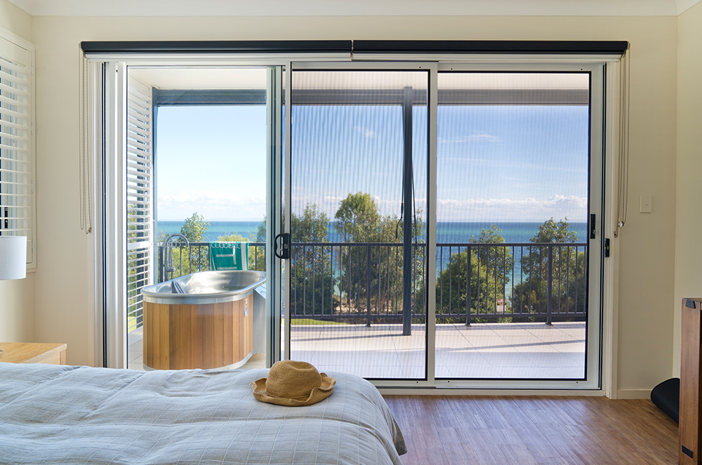Bedroom with sliding glass doors and fitted security screens opening onto a balcony with ocean views, timber flooring, and natural light.